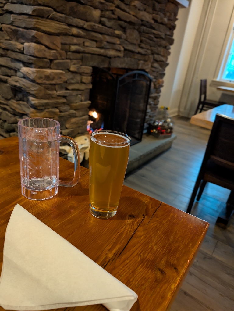 This picture a glass of beer and a glass off water on a table. There is a burning fireplace in the background.