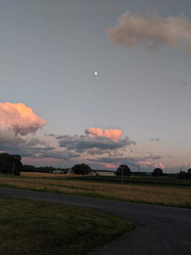 This is a picture of a bright moon in the dusky sky. There are some cumulus clouds below it illuminated from the setting sun.