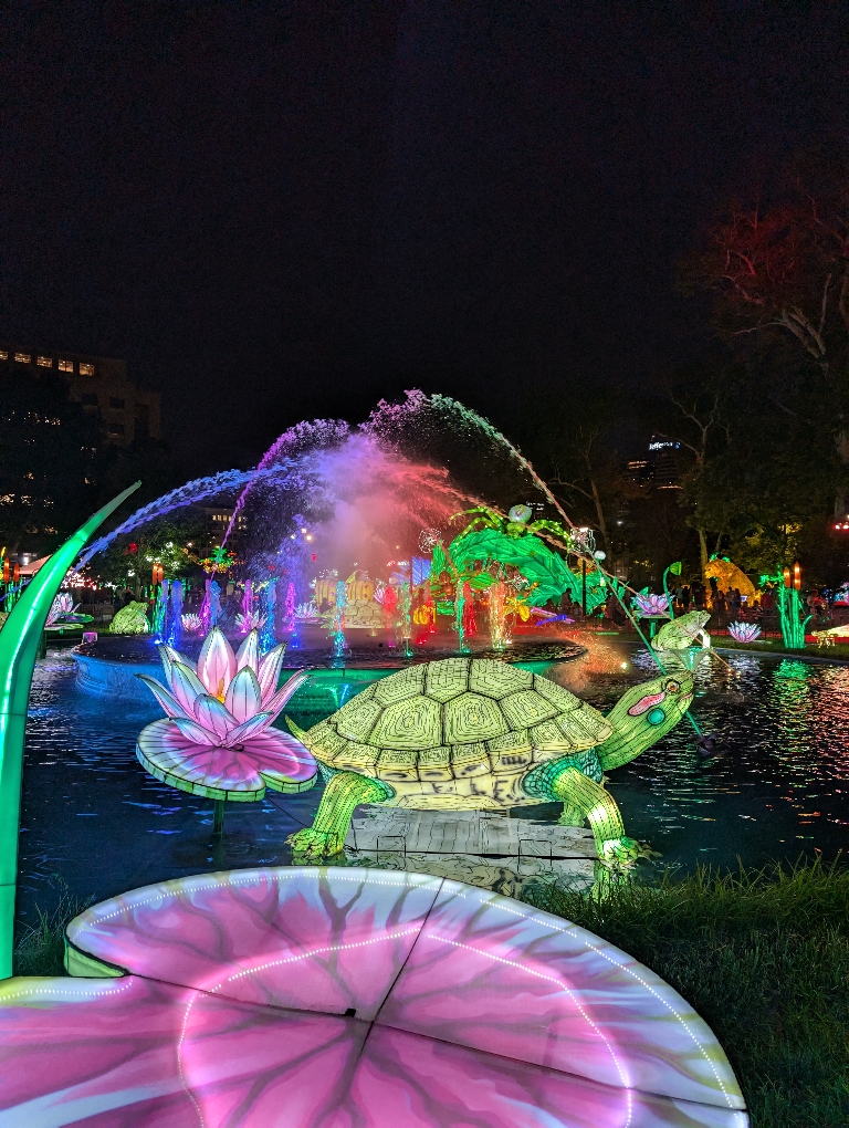 This is picture of the illuminated fountain by all the colors of the rainbow with a green lit up turtle in front of it.