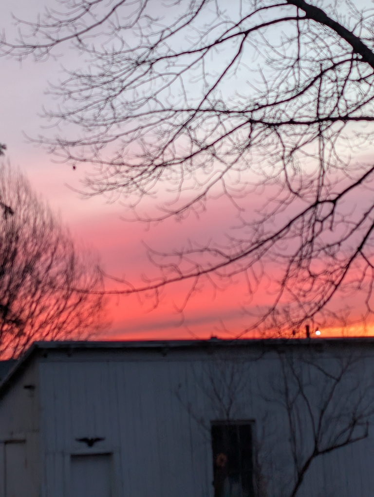 This picture shows a red, orange, pink, and gray horizon from a sunrise. It's just above a garage roof.