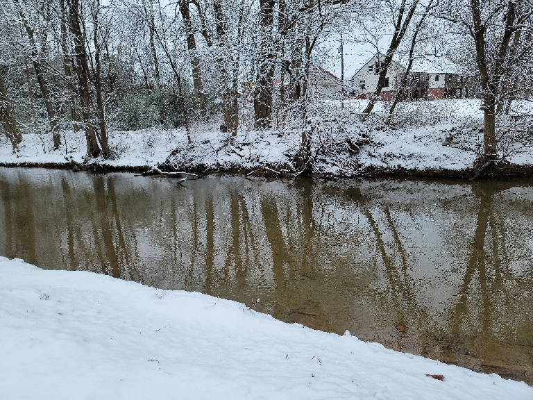 This picture shows the waters of a pretty big creek with wet snow surrounding it including the banks and stuck to the trees all along it.