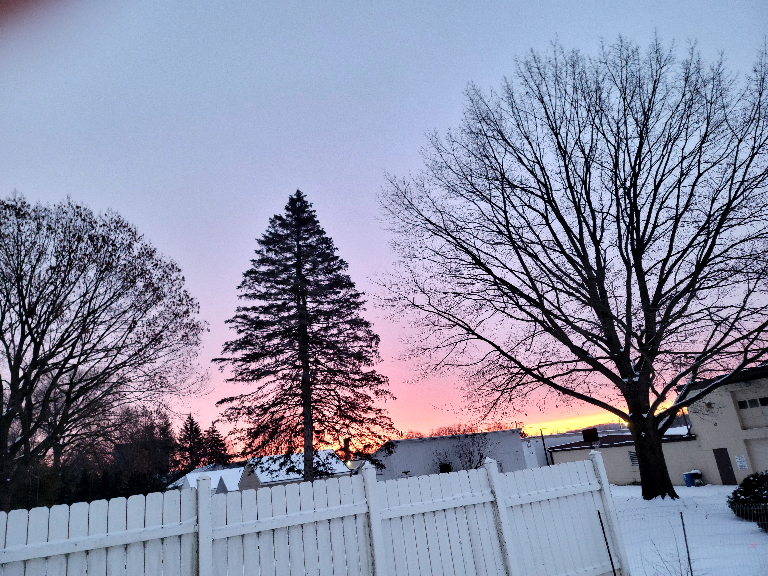 This picture shows a snow covered neigborhood with a nice colorful sunrise in the background.