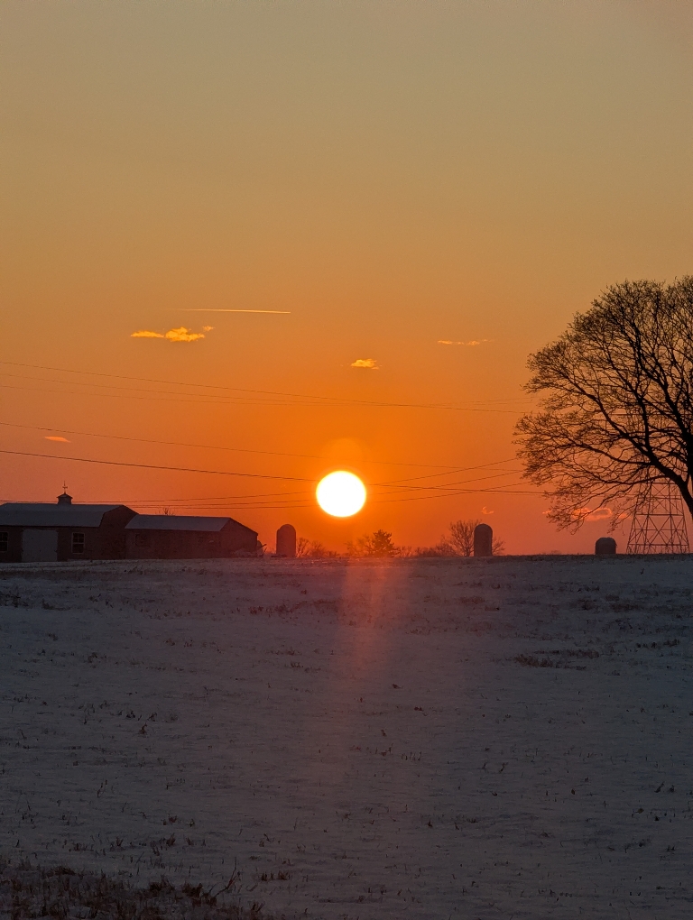 This picture shows an orange setting sun over a snow-covered farm. There are also a few illuminated clouds.