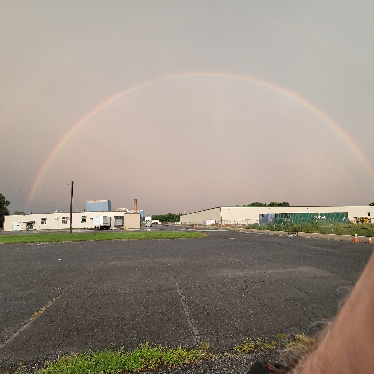 This is a photo of a double rainbow over a small factory type of building near sunset.